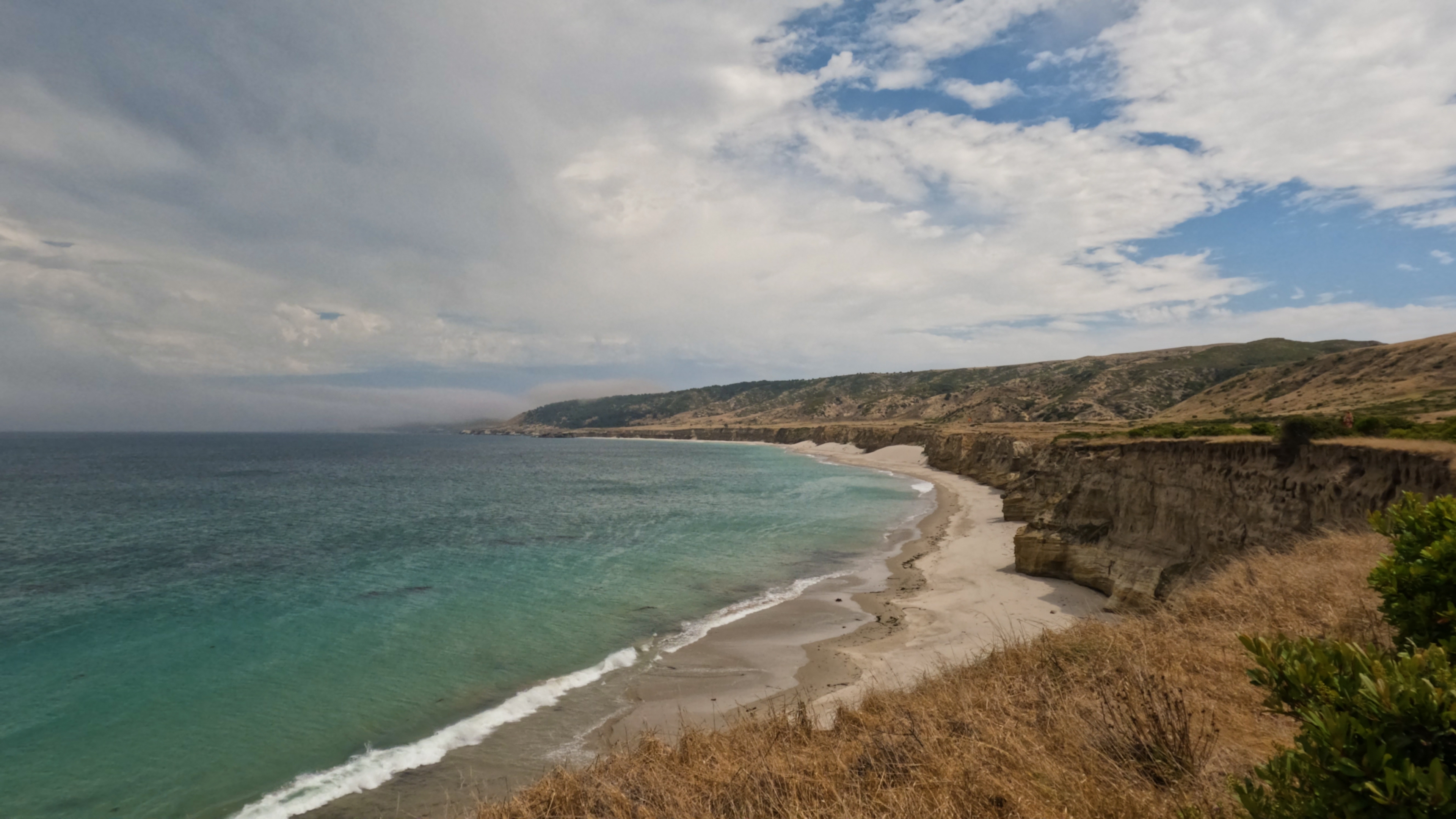 A scenic coastal view featuring rugged cliffs and a sandy beach curving along the shoreline, with turquoise waves gently lapping against the shore. The landscape is covered in dry grass and low vegetation under a partly cloudy sky with patches of blue.