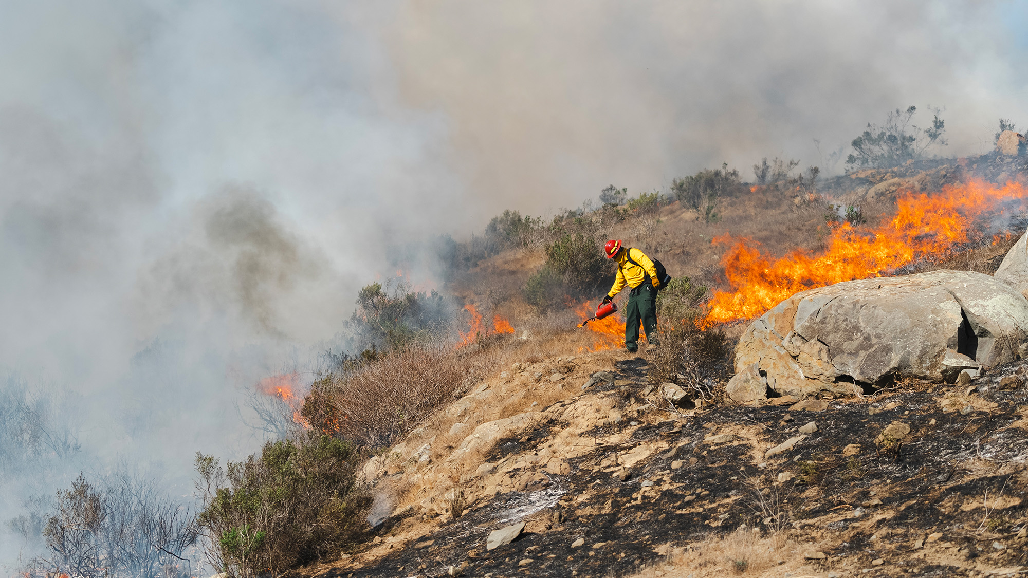 A wildland firefighter in protective gear uses a drip torch to conduct a controlled burn on a dry hillside. Flames spread through brush while thick smoke rises into the air, with large rocks and sparse vegetation visible in the rugged terrain.
