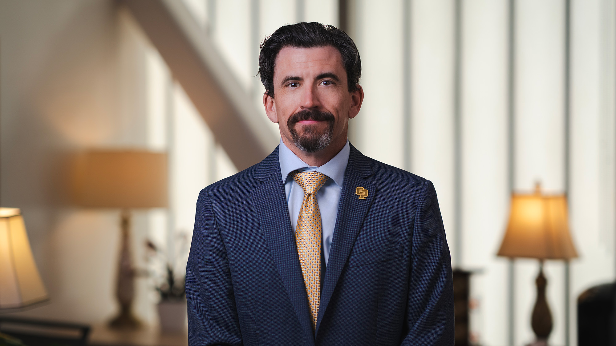 Dean Daniel R. Bernard in a navy suit and gold tie, standing indoors with soft-lit lamps in the background