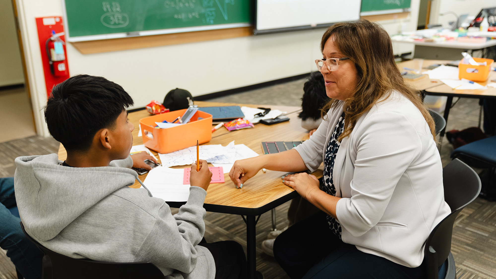 A teacher sits at a classroom table engaging with a middle school student who is holding a pencil and worksheet. The table is scattered with papers, school supplies, and a bright orange supply bin, while a chalkboard and projector screen are visible in the background.