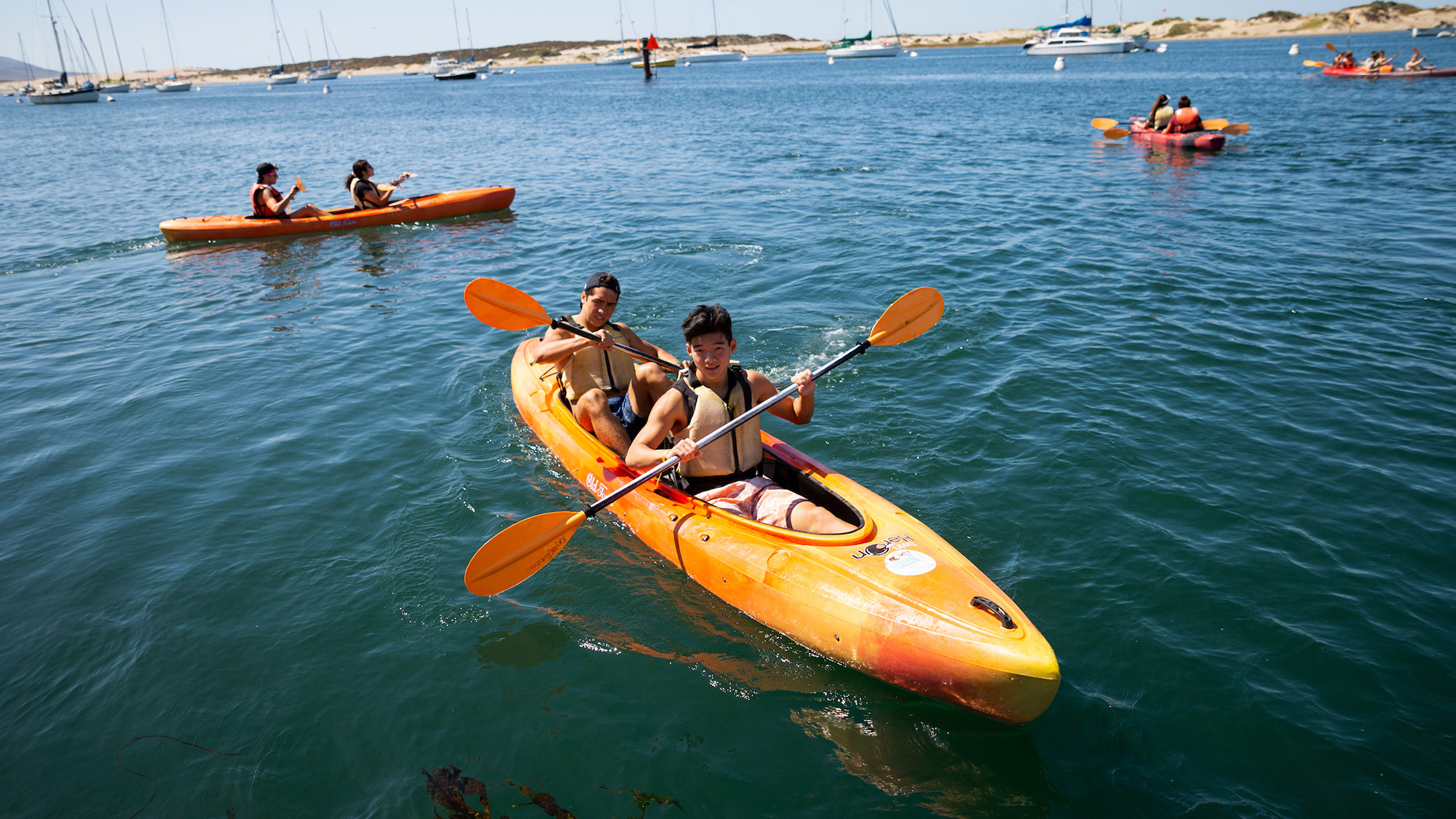 Two students paddle an orange tandem kayak on calm blue water during a Quarter Plus outdoor activity, with other kayakers and sailboats visible in the background under a sunny sky.