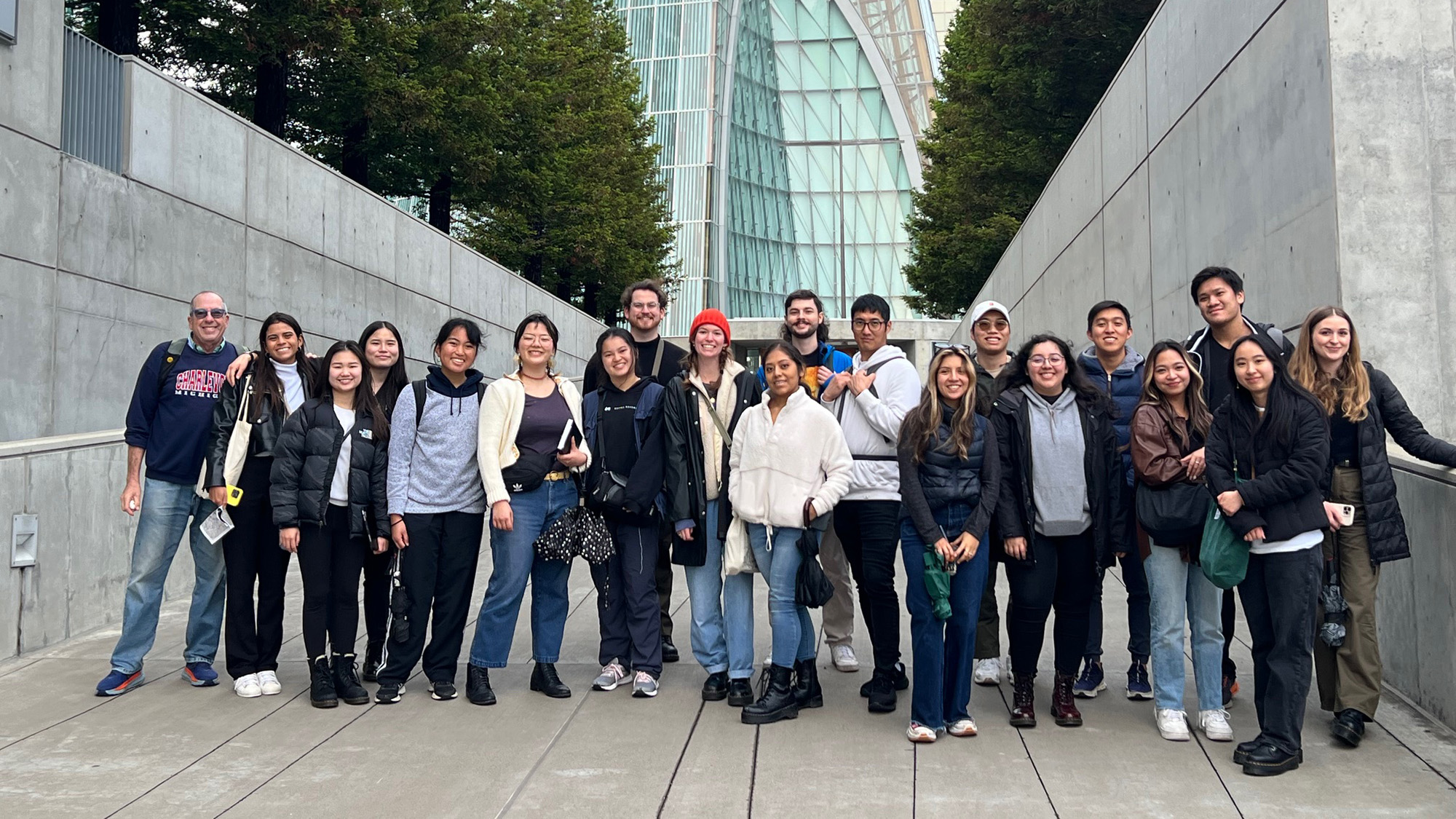 A group of Cal Poly architecture students and a faculty member pose for a photo outside a modern glass and concrete building in San Francisco. The students are smiling and dressed for cool weather, standing between tall concrete walls lined with trees.