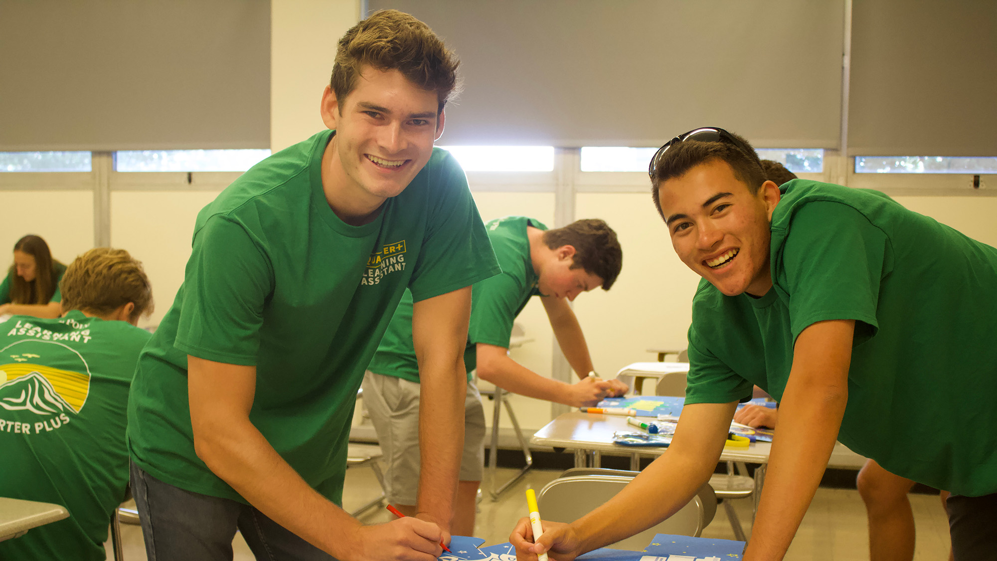Two smiling students in green Cal Poly Quarter Plus shirts lean over desks while working on colorful classroom projects with markers. Other students in the background are also engaged in hands-on activities in a brightly lit classroom.