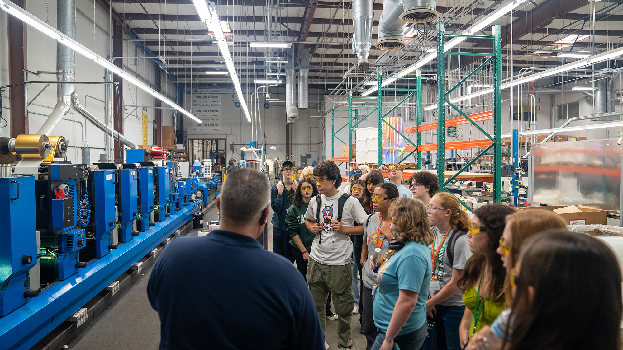 A group of high school students wearing yellow safety glasses listen attentively to a tour guide inside a brightly lit industrial manufacturing facility with blue machinery and exposed ducts. The students appear engaged as they learn about the equipment, with some holding water bottles and lanyards around their necks.