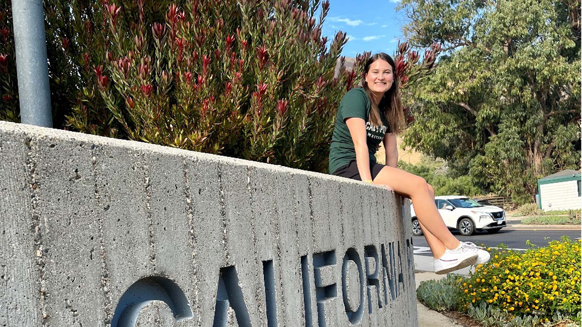 International student sitting on a CALIFORNIA sign at Cal Poly campus, smiling in front of flowers and trees