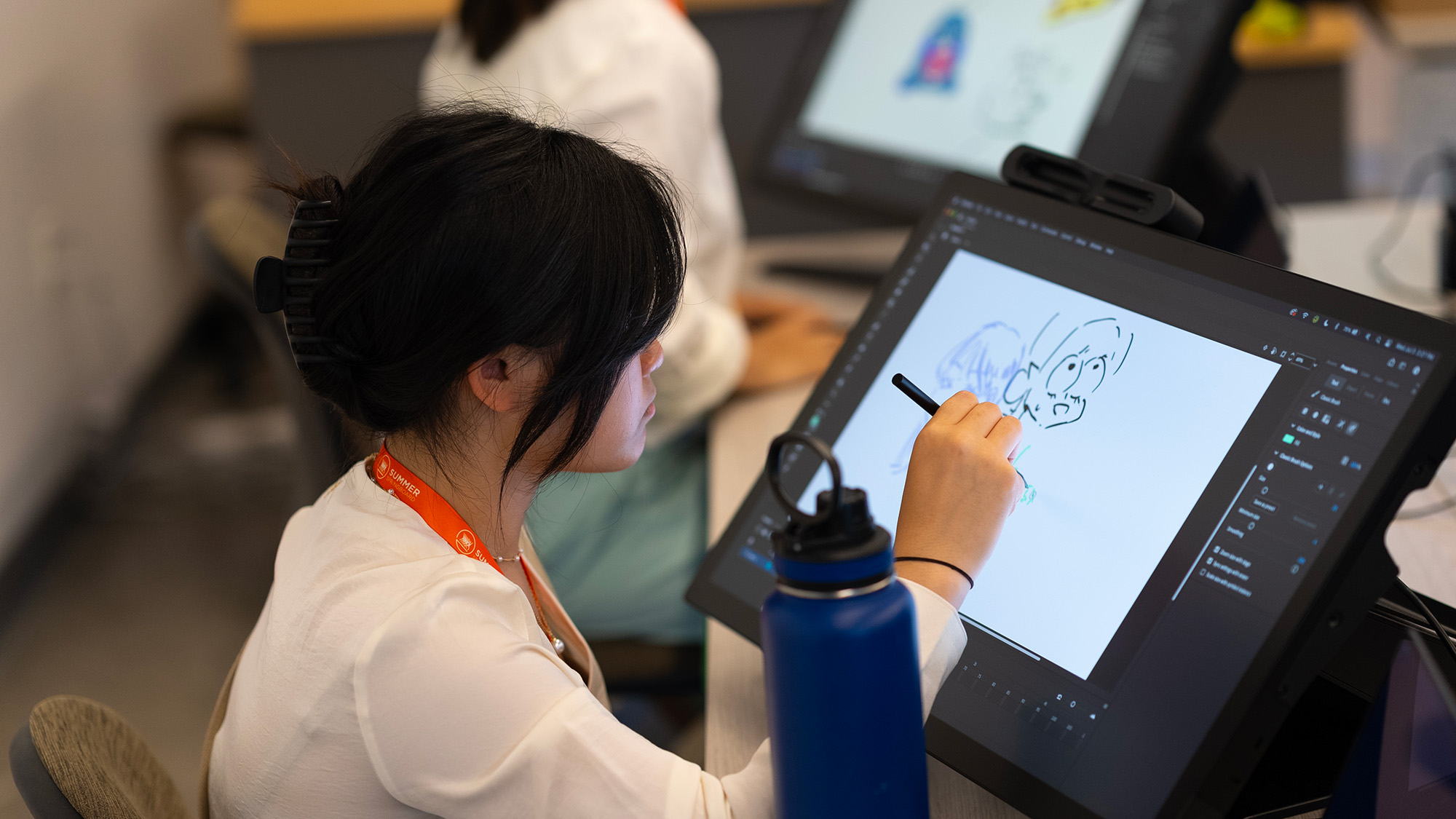 A student drawing a cartoon-style character on a large digital pen tablet during a motion-graphics or animation class, with another student working in the background.